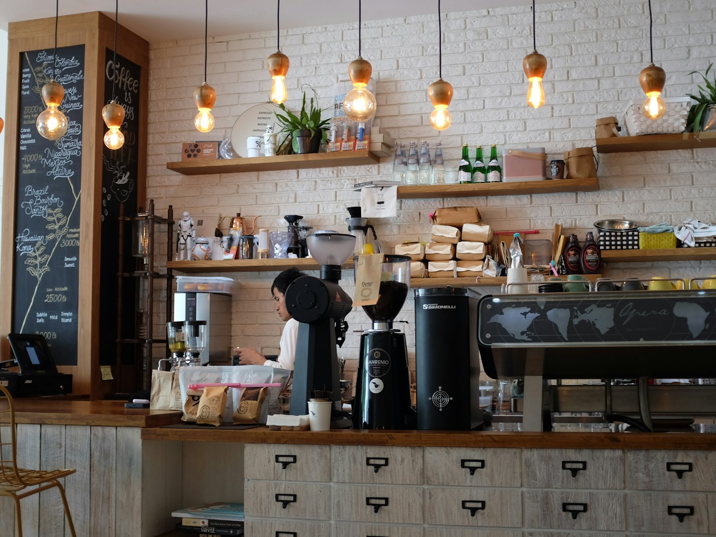 A café counter with an espresso machine, grinders and shelves under pendant lights
